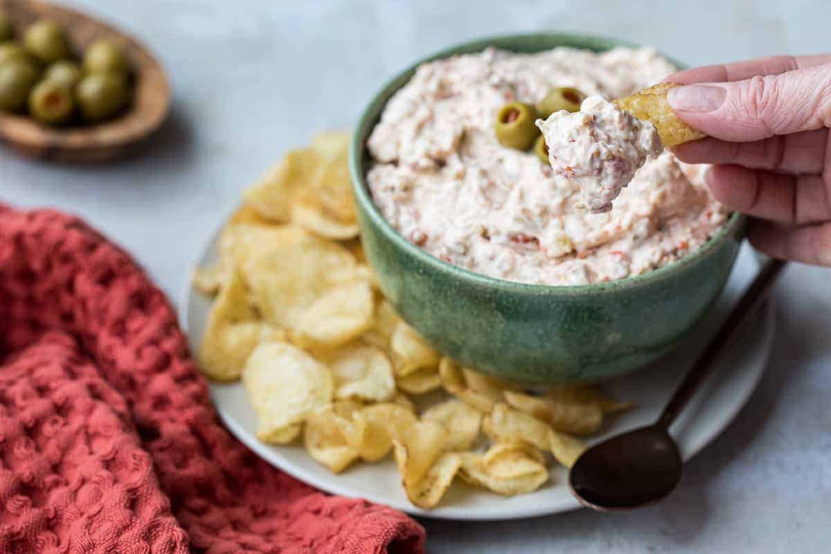 A hand holds a chip with a scoop of creamy dip from a green bowl topped with olives. The bowl is on a plate with more chips, next to a red textured cloth and a small spoon. A small bowl of green olives is in the background.