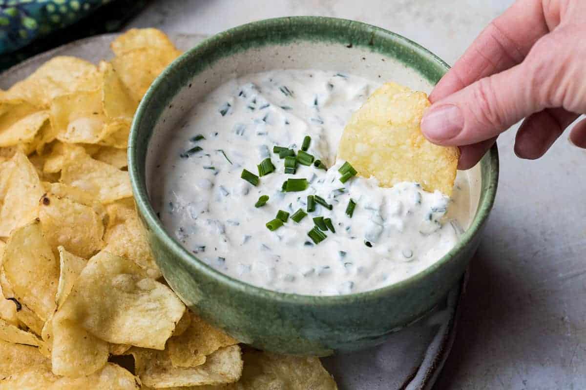 A hand dips a potato chip into a green bowl filled with creamy dip, topped with chopped chives. A pile of potato chips is visible beside the bowl.
