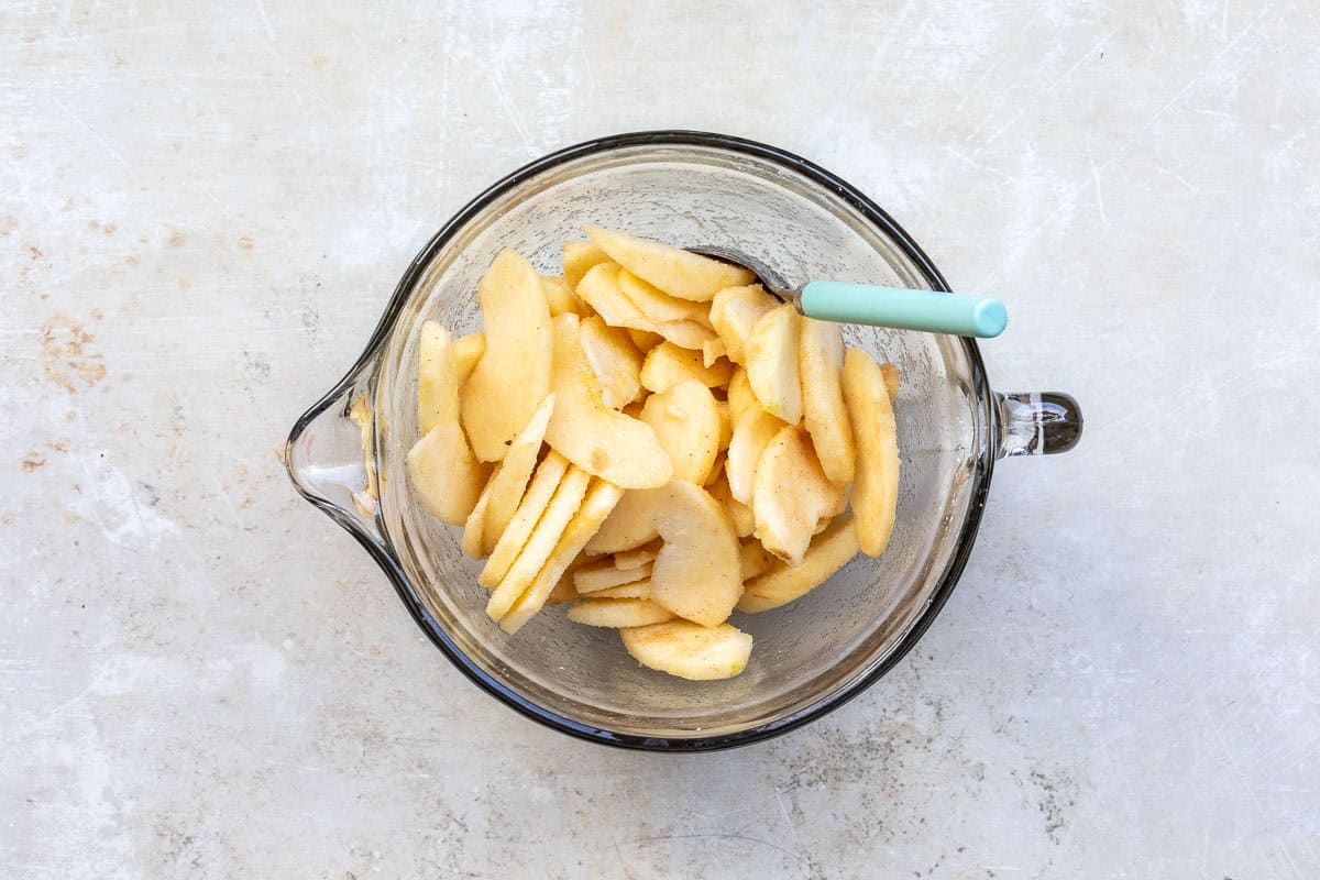 A glass mixing bowl filled with sliced apples, lightly coated, with a teal spoon resting inside. The background is a textured light surface.