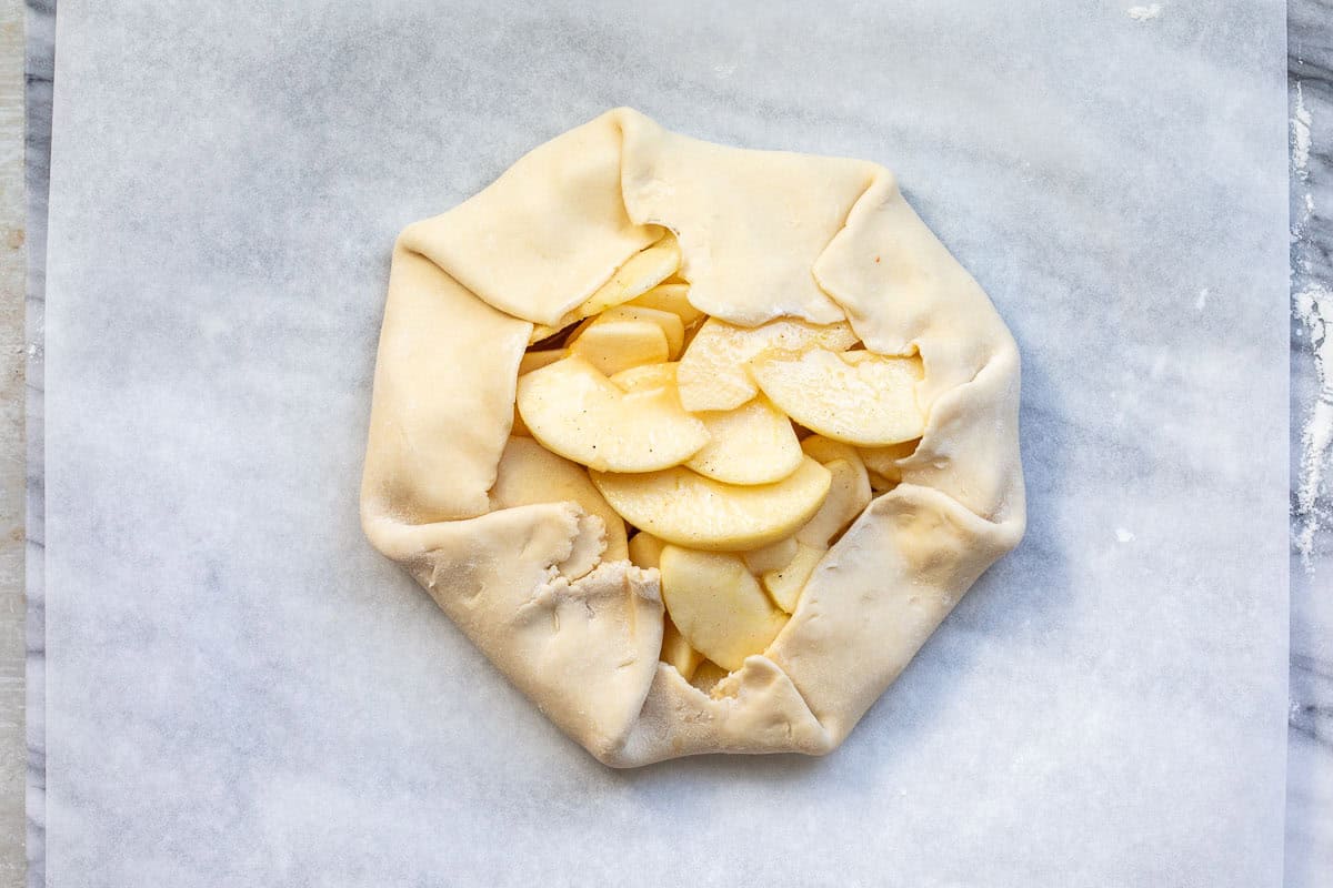 A partially folded apple galette on parchment paper, showing slices of apples stacked in the center of a round pie crust with folded edges. The galette is ready to be baked.