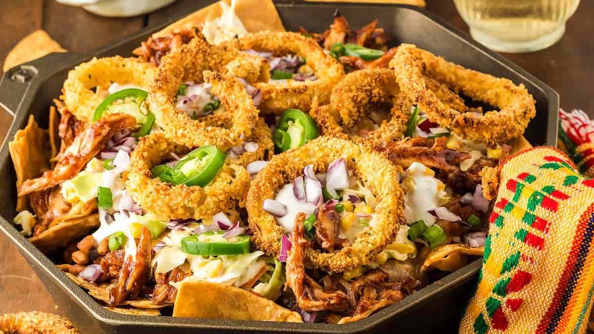 A skillet filled with nachos topped with shredded meat, jalapeño slices, diced onions, crispy onion rings, and melted cheese. A colorful fabric napkin is partially visible on the side.