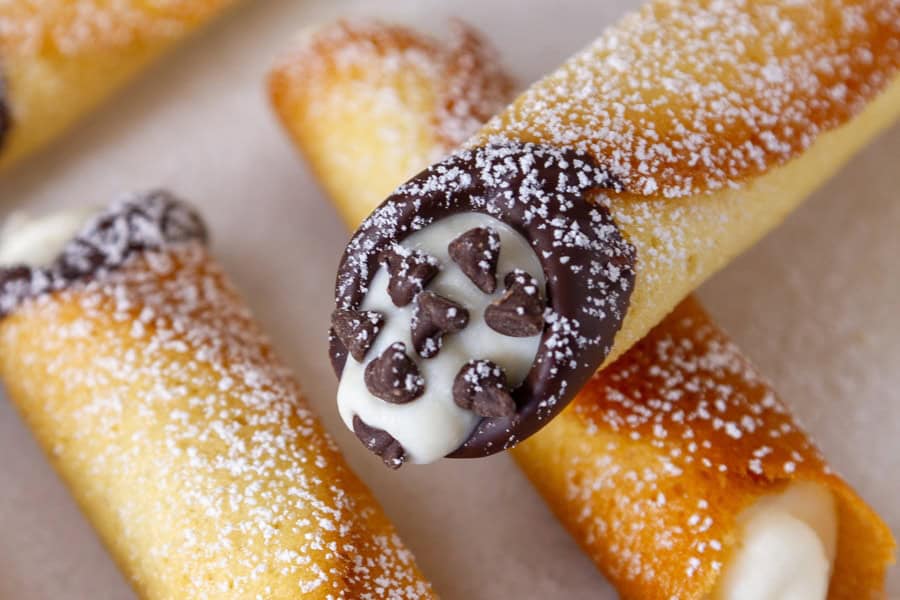 Close-up of cream-filled cannoli dusted with powdered sugar, perfect for a Valentines Day treat. The ends are dipped in chocolate and topped with small chocolate chips, placed on a light-colored surface.
