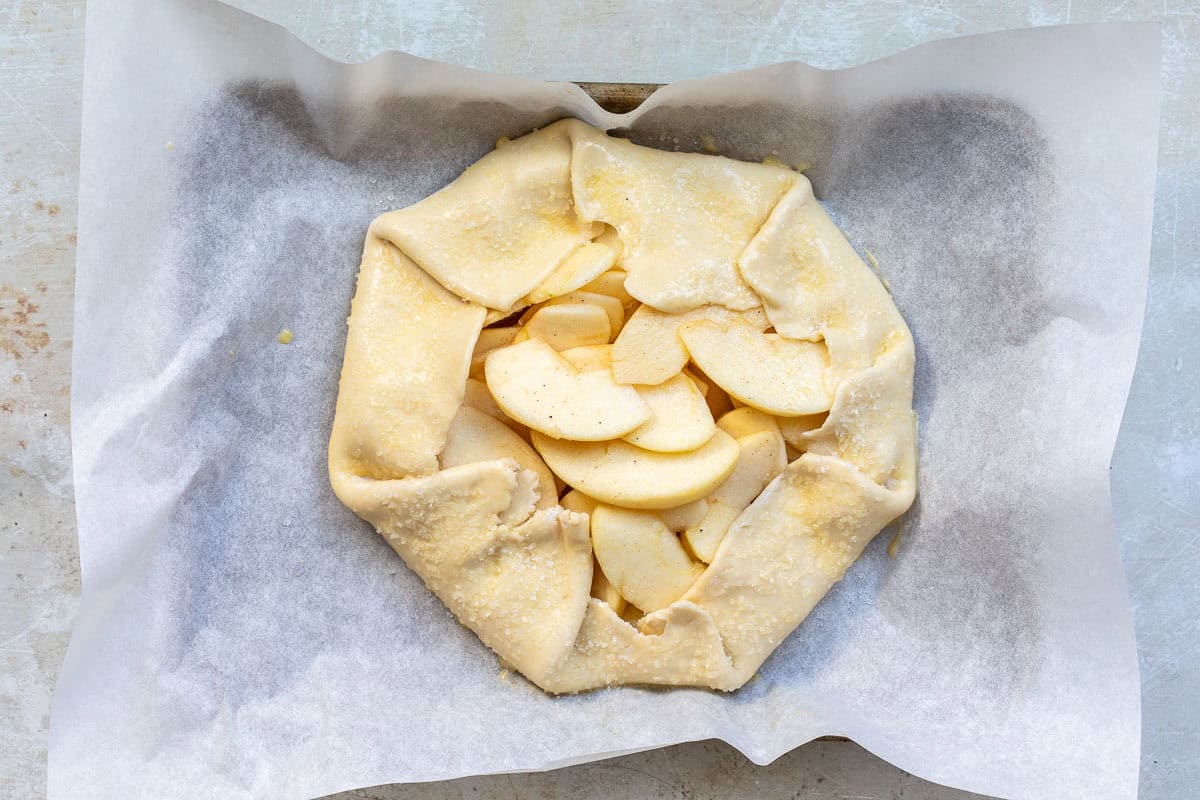 A rustic, unbaked apple galette with folded edges, filled with sliced apples on parchment paper. The crust is sprinkled with sugar, and the galette is on a baking tray ready to be baked.