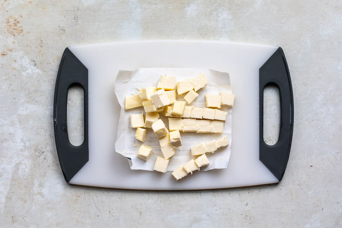 Diced cubes of butter on a sheet of parchment paper placed on a white cutting board with black handles, viewed from above. The surrounding surface is a light, textured countertop.