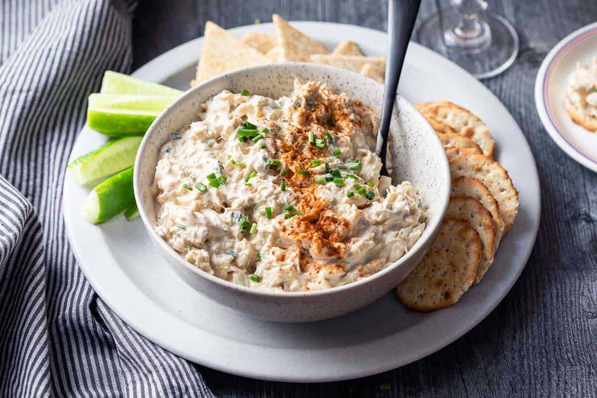 A bowl of creamy dip topped with paprika and chopped chives, surrounded by crackers, lime wedges, and pita bread on a plate. A spoon is in the bowl, and a striped cloth napkin is to the left.
