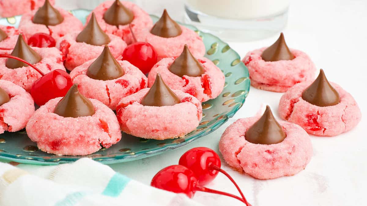 A green plate holds several pink Valentines Day cookies with chocolate kisses on top, adorned with cherry bits. A few cherries are scattered next to the plate, and a glass of milk is visible in the background.