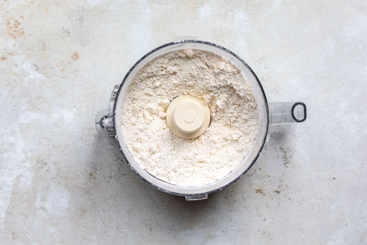 A food processor bowl filled with a flour/butter mixture, sitting on a textured, light-colored surface. The processor lid is off, showing a central plastic blade.