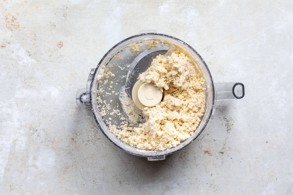 A food processor bowl filled with crumbly pale dough on a light, textured surface. The pie dough is rough and unevenly distributed around the central blade.
