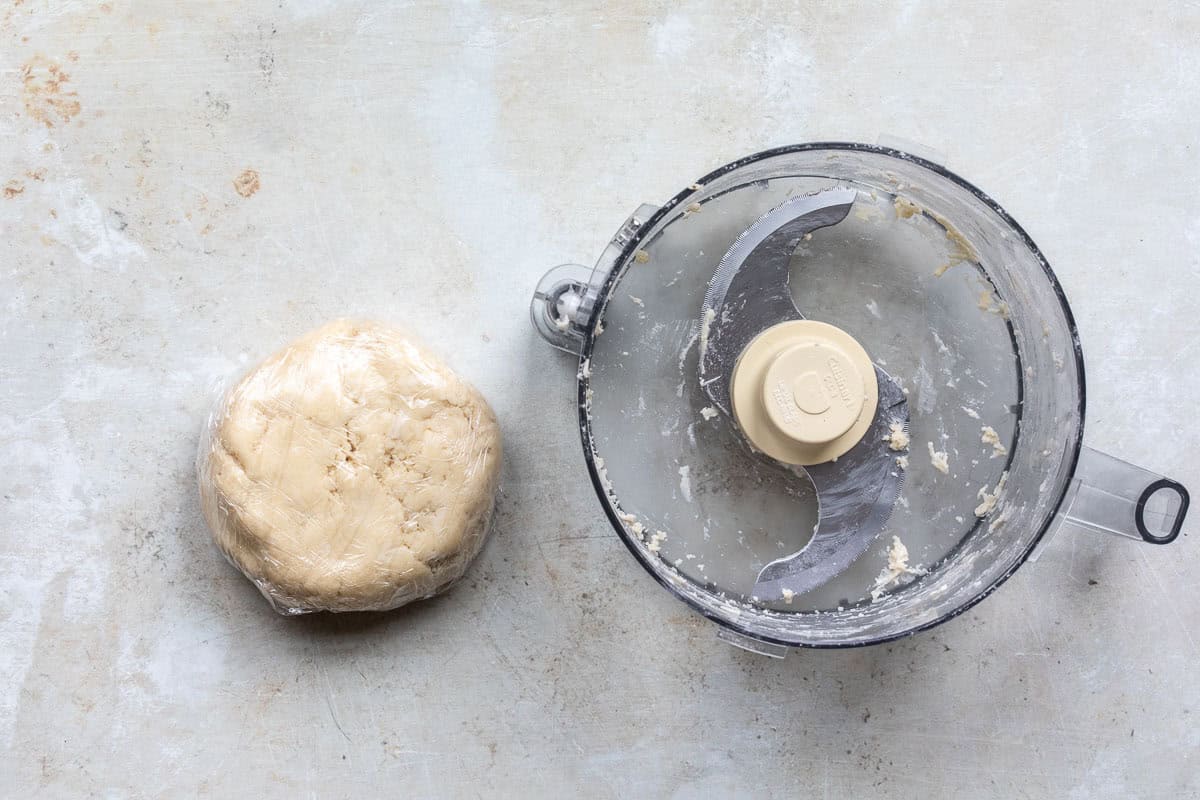 A ball of dough wrapped in plastic sits next to an empty food processor with dough residue inside, on a light gray surface.