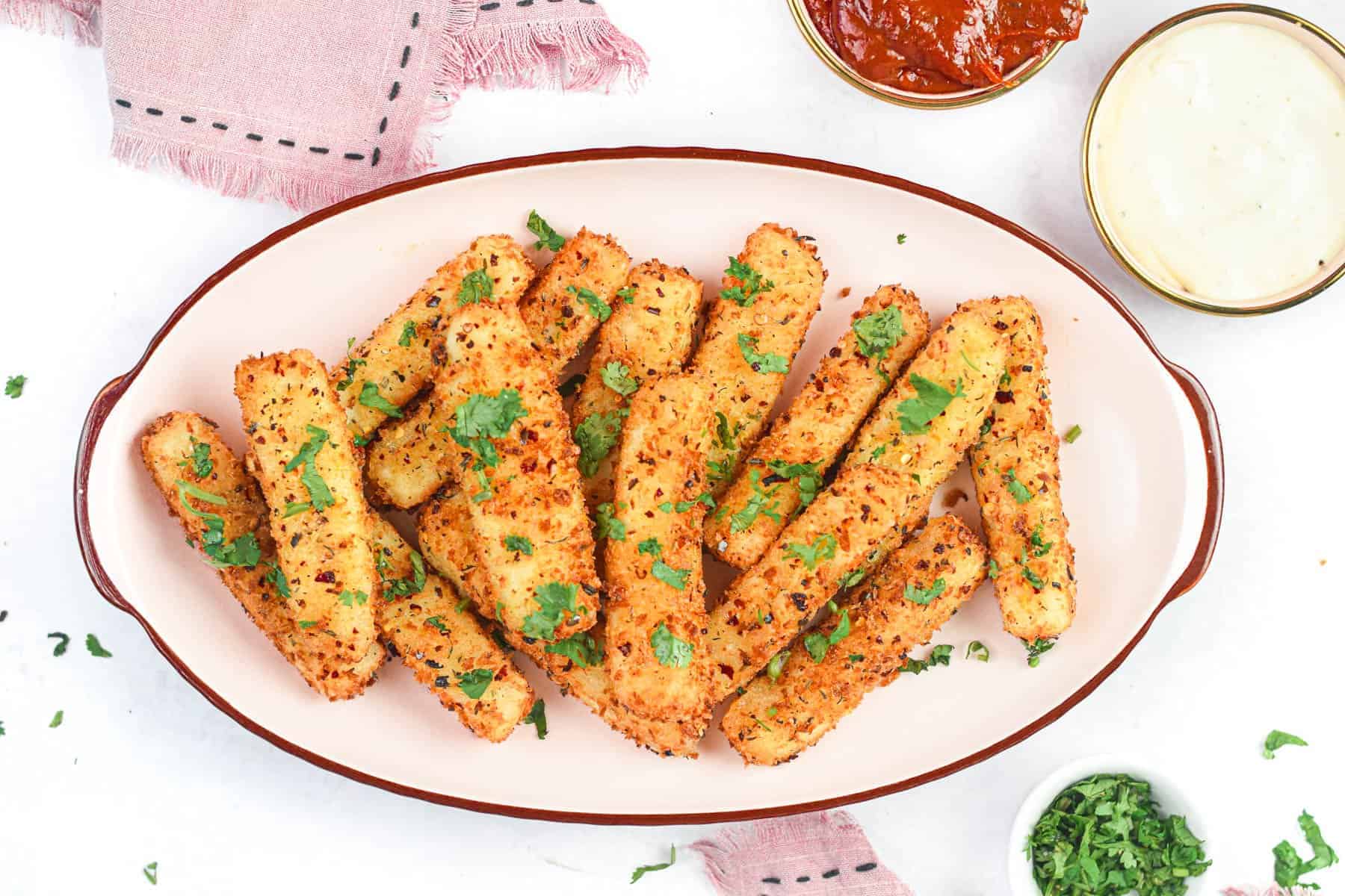 A plate of crispy golden zucchini fries garnished with parsley. Two small bowls of red and white dipping sauces are nearby on a pink cloth. Additional chopped parsley is in a bowl to the side.