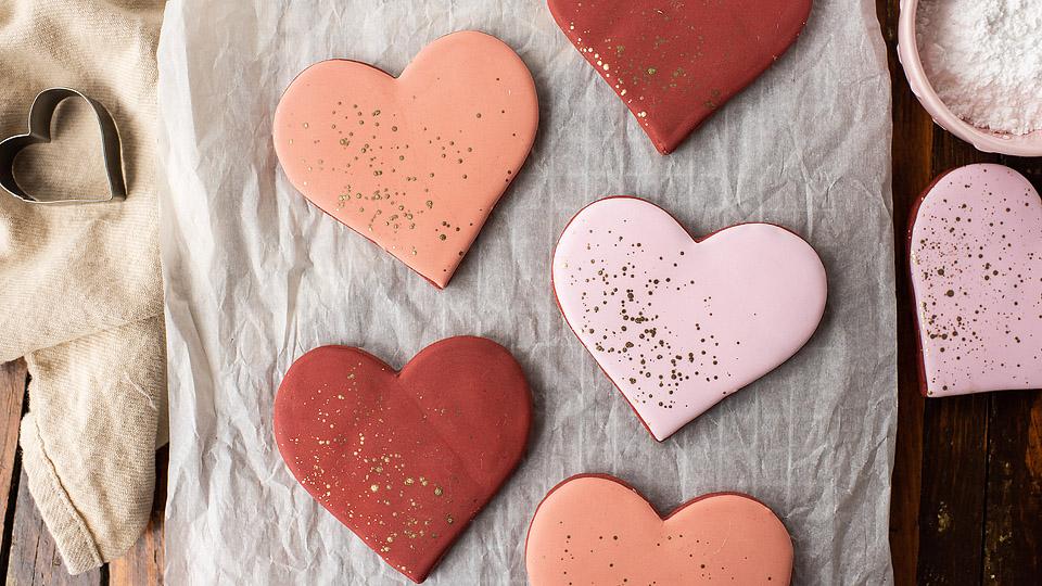 Heart-shaped cookies with pink and red icing are arranged on parchment paper, perfect for Valentines Day. A dusting of gold sprinkles decorates each cookie, capturing the festive spirit. Nearby, a small heart-shaped cookie cutter and a bowl of powdered sugar add to the sweet scene.