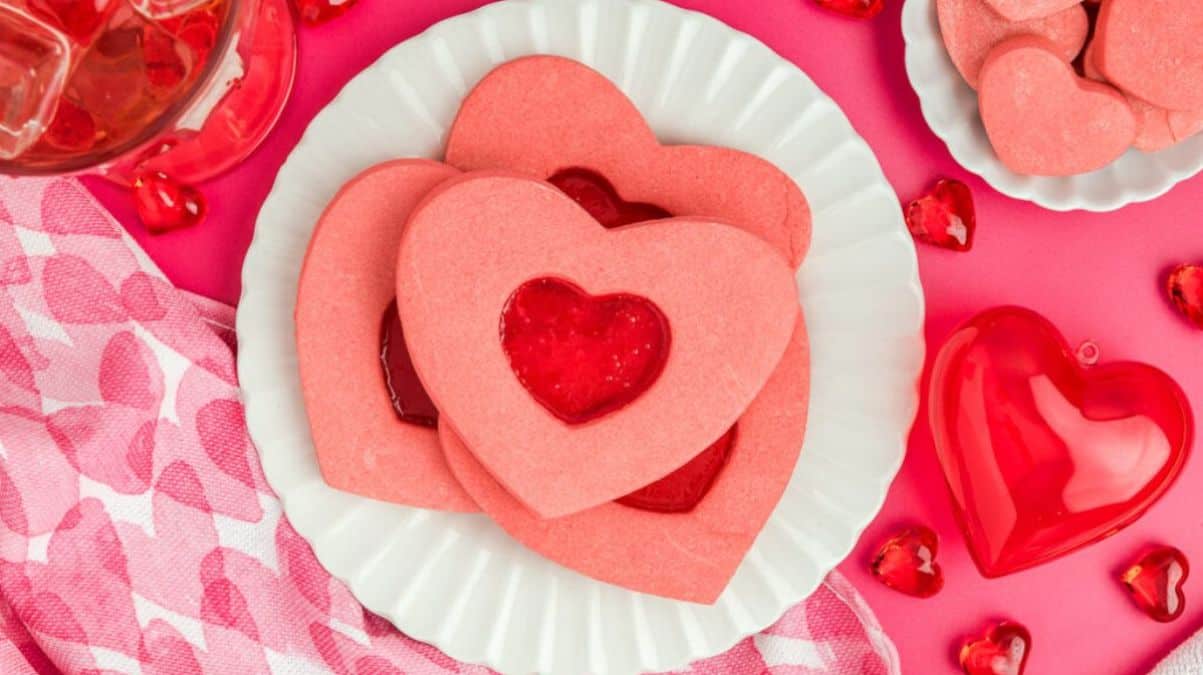 Heart-shaped pink Valentines Day cookies with a red jam center are arranged on a white plate. Set against a pink background, a pink and white checkered cloth lies nearby. Small decorative red heart ornaments surround the plate.