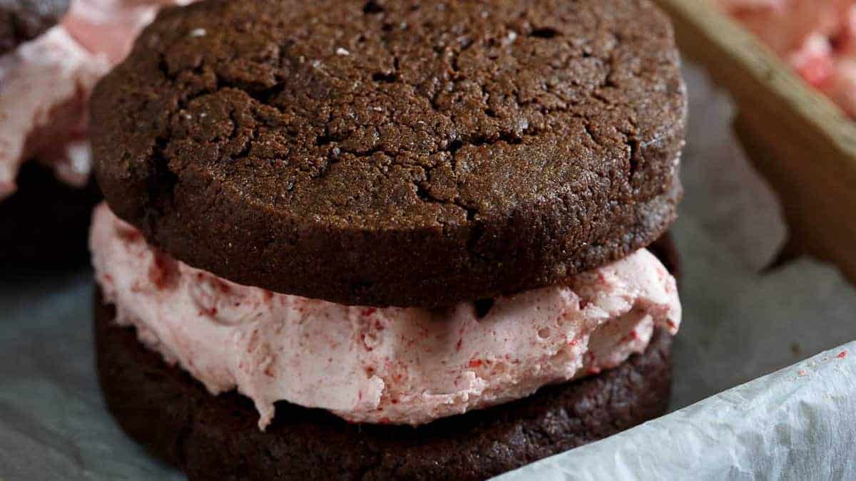 Two Valentines Day cookies sandwich a generous layer of pink strawberry ice cream, sitting on wax paper. The ice cream has small chunks, adding texture to the dessert.