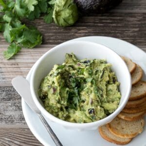 A bowl of roasted tomatillo guacamole on a plate with crostini. Fresh cilantro and an avocado are in the background on a wooden surface.