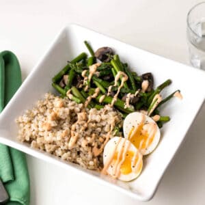 A square bowl contains pearl barley, asparagus, mushrooms, and two halved boiled eggs, drizzled with sauce. A green napkin and a glass of water are nearby.