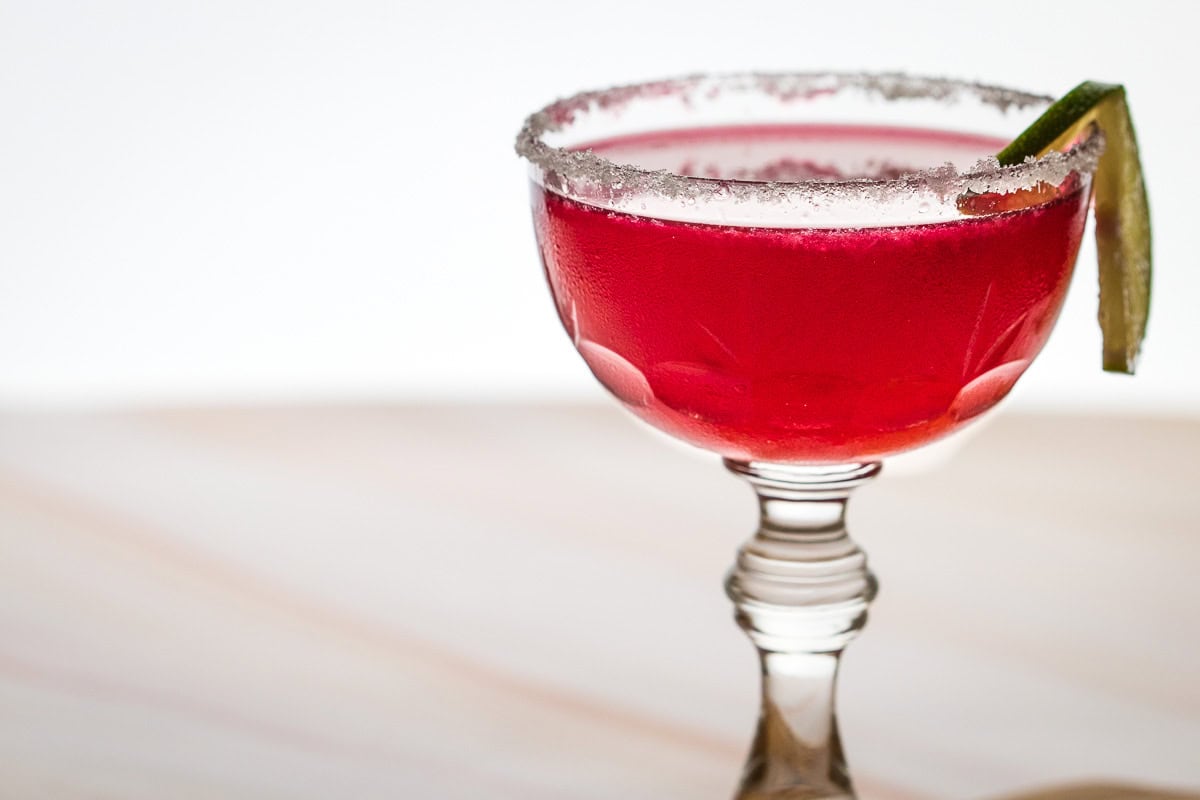A glass of hibiscus margarita cocktail with a sugared rim and a lime wedge, set on a wooden surface.