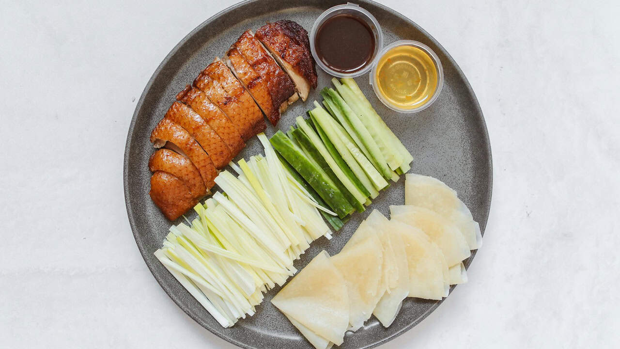 A plate with sliced roast duck, cucumber sticks, julienned scallions, thin pancakes, and two small containers of sauce—an iconic dish every foodie should try—arranged neatly on a gray dish.