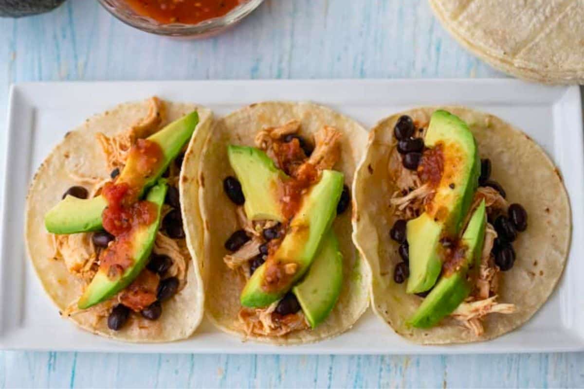 Three tacos on a white plate topped with shredded chicken, black beans, avocado slices, and salsa—perfect for busy weeknights when you need easy chicken dinners the whole family will love. This dish is a delicious addition to your collection of chicken recipes.