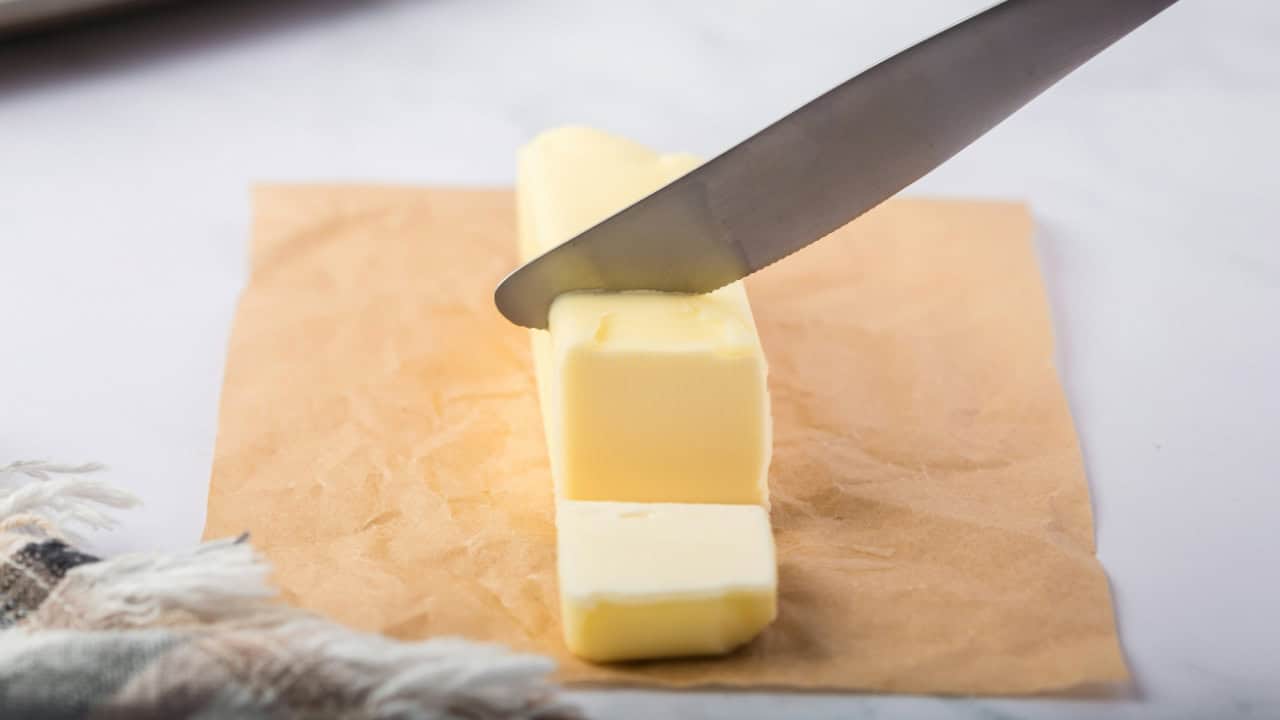 A Caucasian hand holds a knife slicing a stick of butter which sits on parchment paper with a linen napkin nearby and a baking sheet with cookies in the background