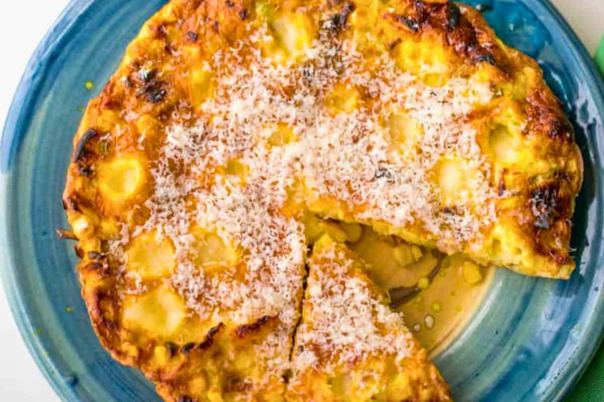 A golden-brown frittata—perfect for summer recipes—topped with grated cheese sits on a blue plate, with one triangular slice cut out from the whole.