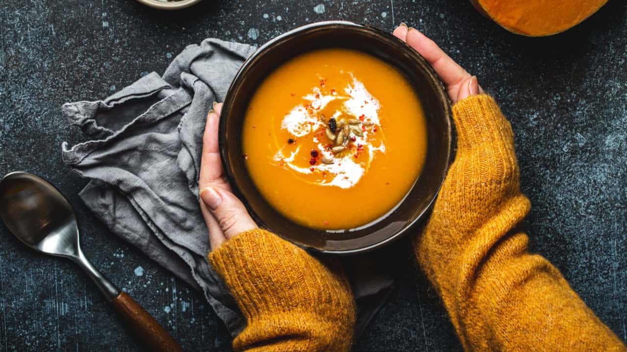 Female hands in yellow knitted sweater holding a bowl with pumpkin cream soup on dark stone background with spoon decorated with cut fresh pumpkin, top view. Autumn cozy dinner concept