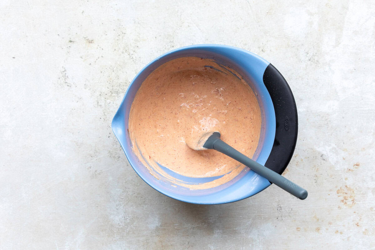 A blue mixing bowl with a black handle contains homemade harissa aioli and a gray spatula rests inside, placed on a light-colored surface.
