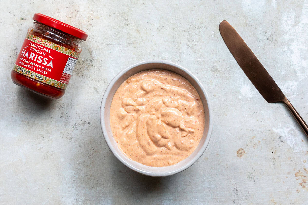 A bowl of creamy harissa sauce sits next to a jar of harissa paste and a brown-handled butter knife on a light-colored surface.