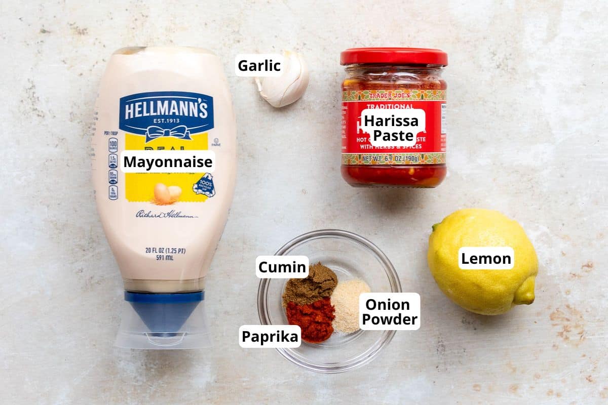 A bottle of mayonnaise, a jar of harissa paste, a garlic clove, a lemon, and a bowl containing cumin, paprika, and onion powder on a light surface.