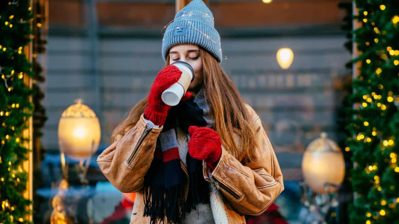 Hipster woman traveler drinking coffee from disposable cup outdoor over beautyful cozy shop window with Christmas decoration.