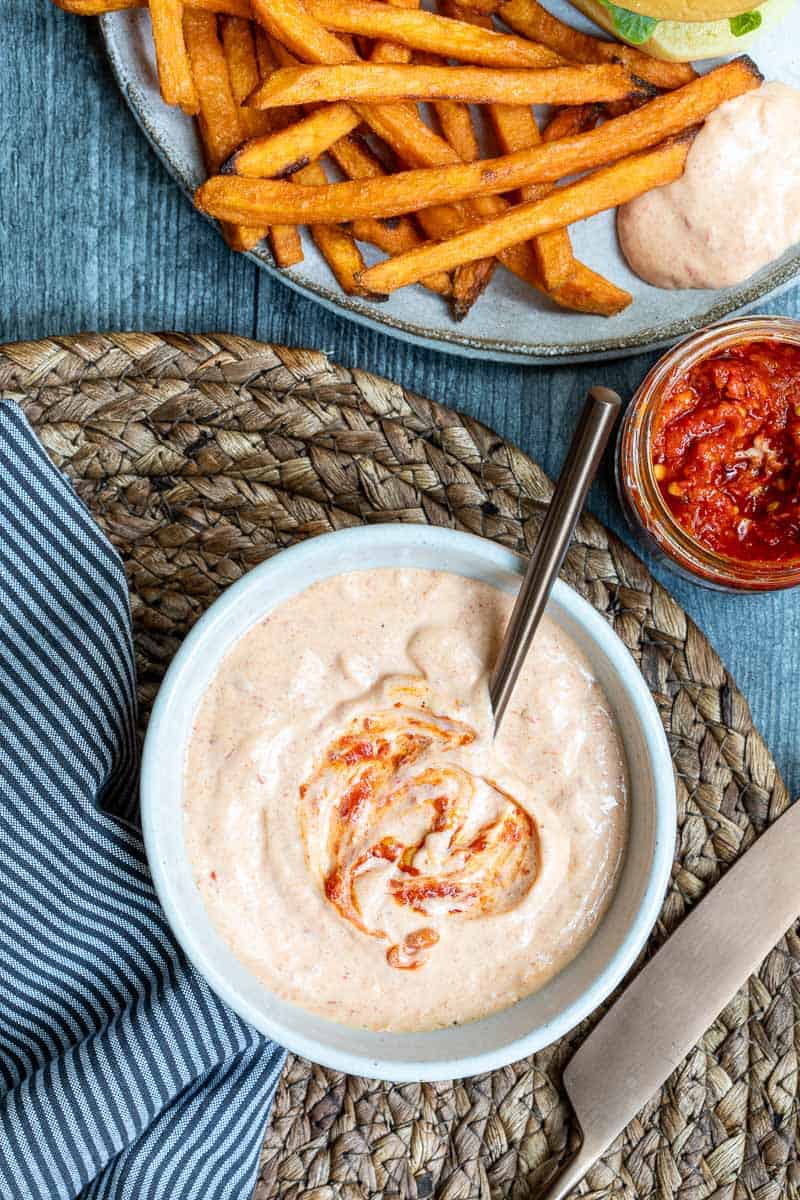 A bowl of creamy harissa aioli sauce with a swirl of harissa, a spoon inside, a plate of fries, and a jar of harissa on a blue table.