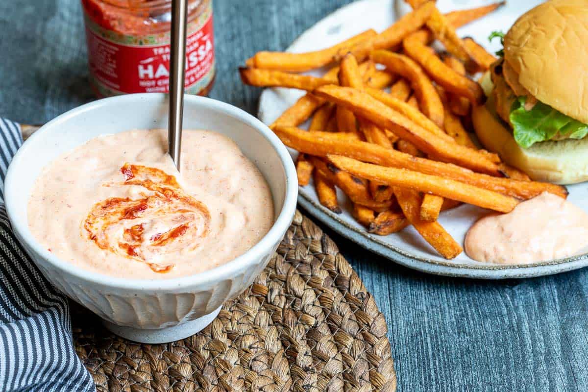 A bowl of creamy harissa aioli with a swirl of harissa sits next to a plate of sweet potato fries and a sandwich with lettuce on a bun.