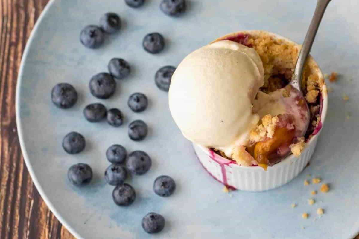 A small ramekin with fruit crumble and a scoop of vanilla ice cream, on a blue plate with scattered blueberries and a spoon—a delightful treat perfect for fans of blueberry desserts.