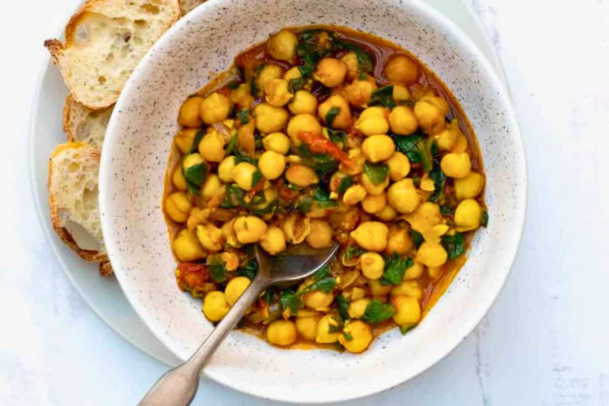 A bowl of chickpea stew with chopped greens and tomatoes, served with sliced bread on a white plate; a spoon is in the bowl. Perfect for fans of healthy meals, this dish is one of those satisfying Chickpea recipes that even meat eaters will love.