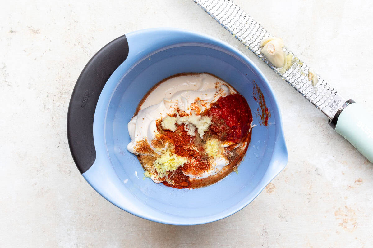 A blue bowl contains mayonnaise, harissa paste, spices, lemon juice, and minced garlic with a grater holding garlic placed beside it on a light-colored surface.