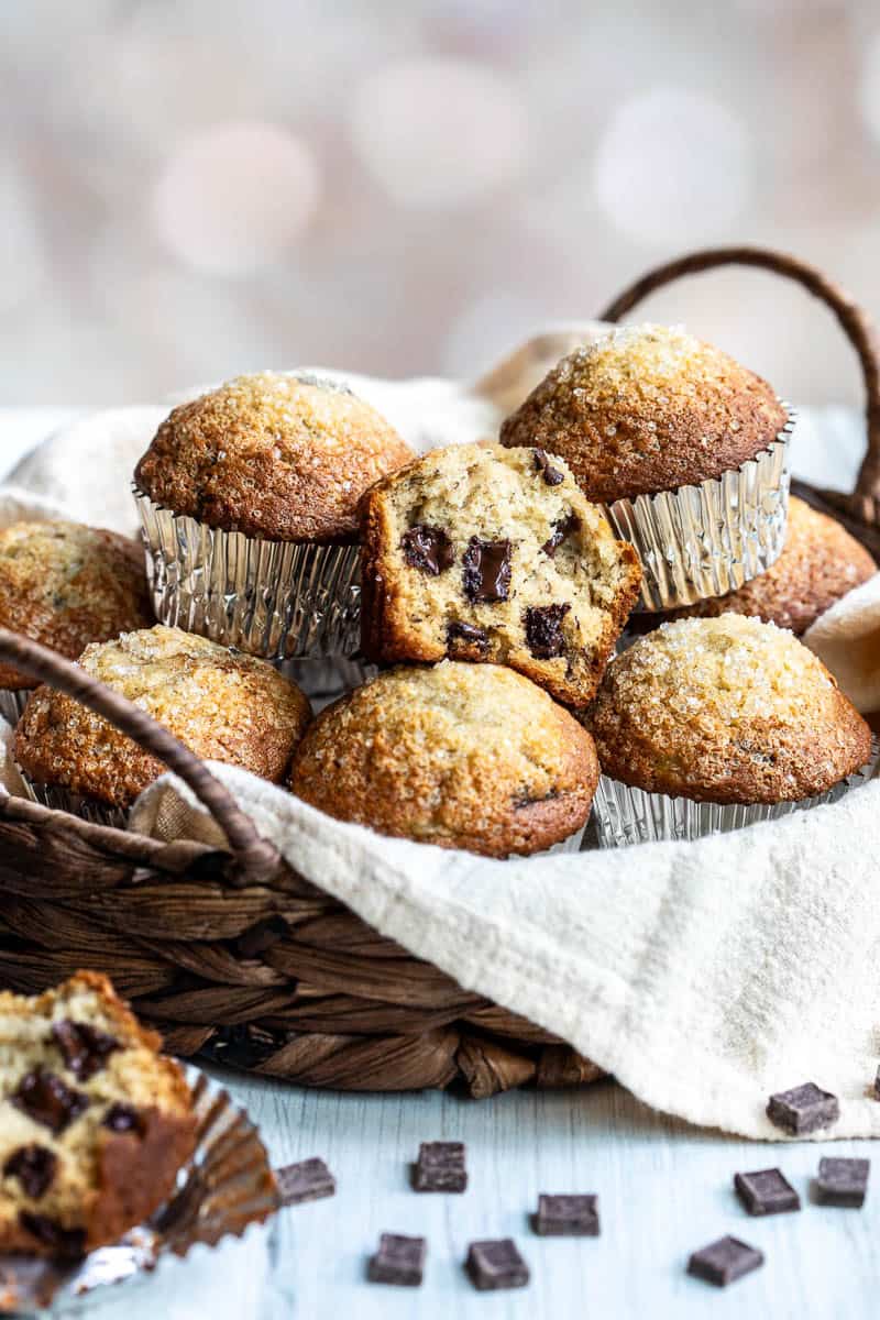 A wicker basket filled with chocolate chunk banana muffins in silver liners, with one muffin cut open to show the chocolate chips inside. Sugar crystals are visible on top of the muffins.