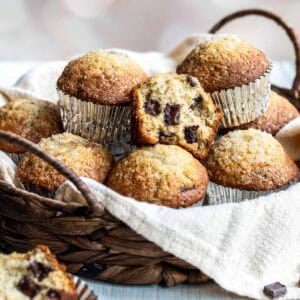 A wicker basket lined with cloth holds several chocolate chunk banana muffins in foil wrappers, with one muffin partially eaten to reveal chunks of chocolate inside.