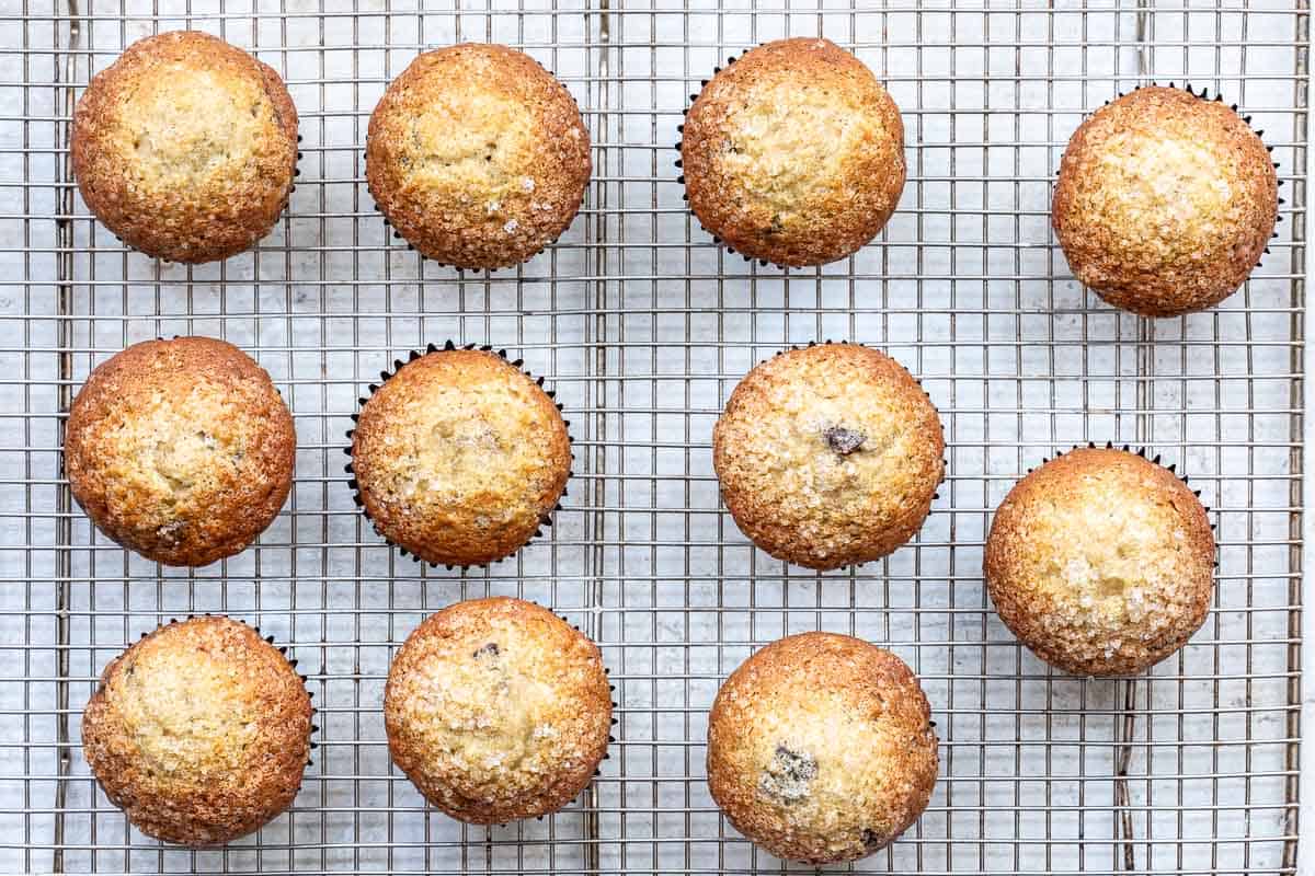 Twelve golden brown banana chocolate chunk muffins with sugar topping are cooling on a wire rack, evenly spaced in three rows of four.