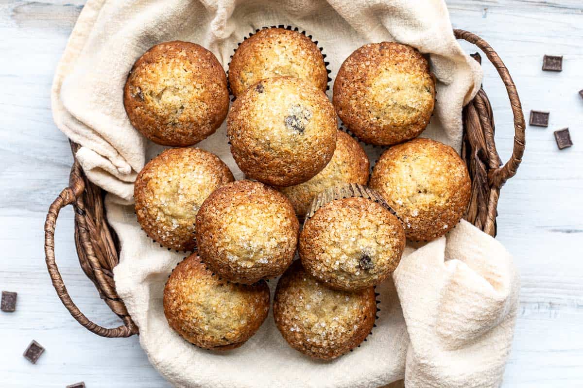 A wicker basket lined with a beige cloth holds twelve golden-brown chocolate chip banana muffins topped with coarse sugar crystals, set on a white wooden surface.