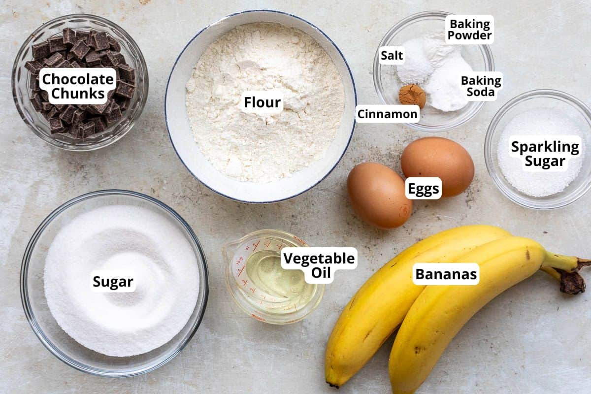 Top view of labeled ingredients for baking banana muffins, including flour, chocolate chunks, sugar, vegetable oil, eggs, bananas, cinnamon, salt, baking soda, baking powder, and sparkling sugar.