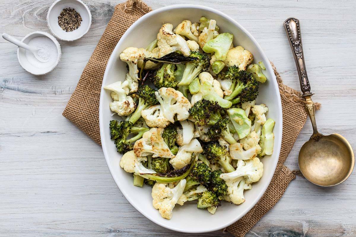 A white bowl filled with roasted cauliflower and broccoli sits on a brown cloth, next to a gold serving spoon and small bowls of salt and pepper on a light wooden table.