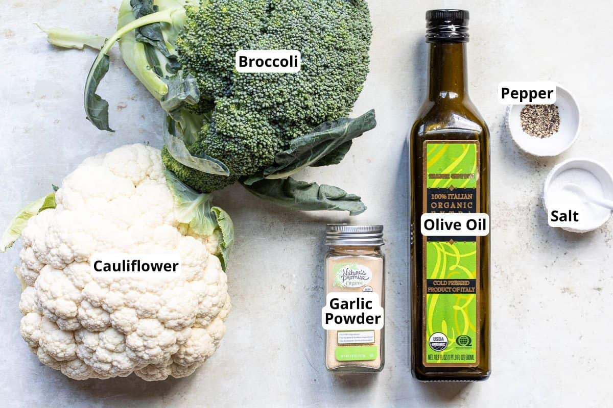 Labeled ingredients on a counter: a head of cauliflower, a head of broccoli, a jar of garlic powder, a bottle of olive oil, and small bowls of salt and pepper.