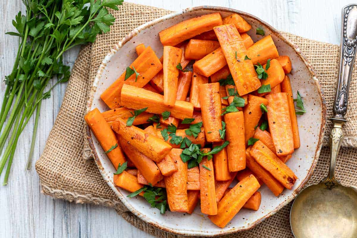 A plate of roasted carrot sticks garnished with chopped parsley, placed on a burlap cloth with fresh parsley and a serving spoon nearby.