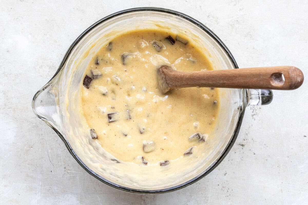 A glass mixing bowl filled with chocolate chunk banana muffin batter and a wooden spoon resting inside.