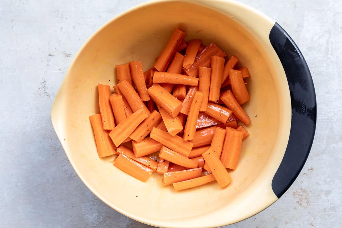 A large yellow mixing bowl filled with peeled, cut carrot sticks coated in oil and seasoning sits on a light-colored surface.