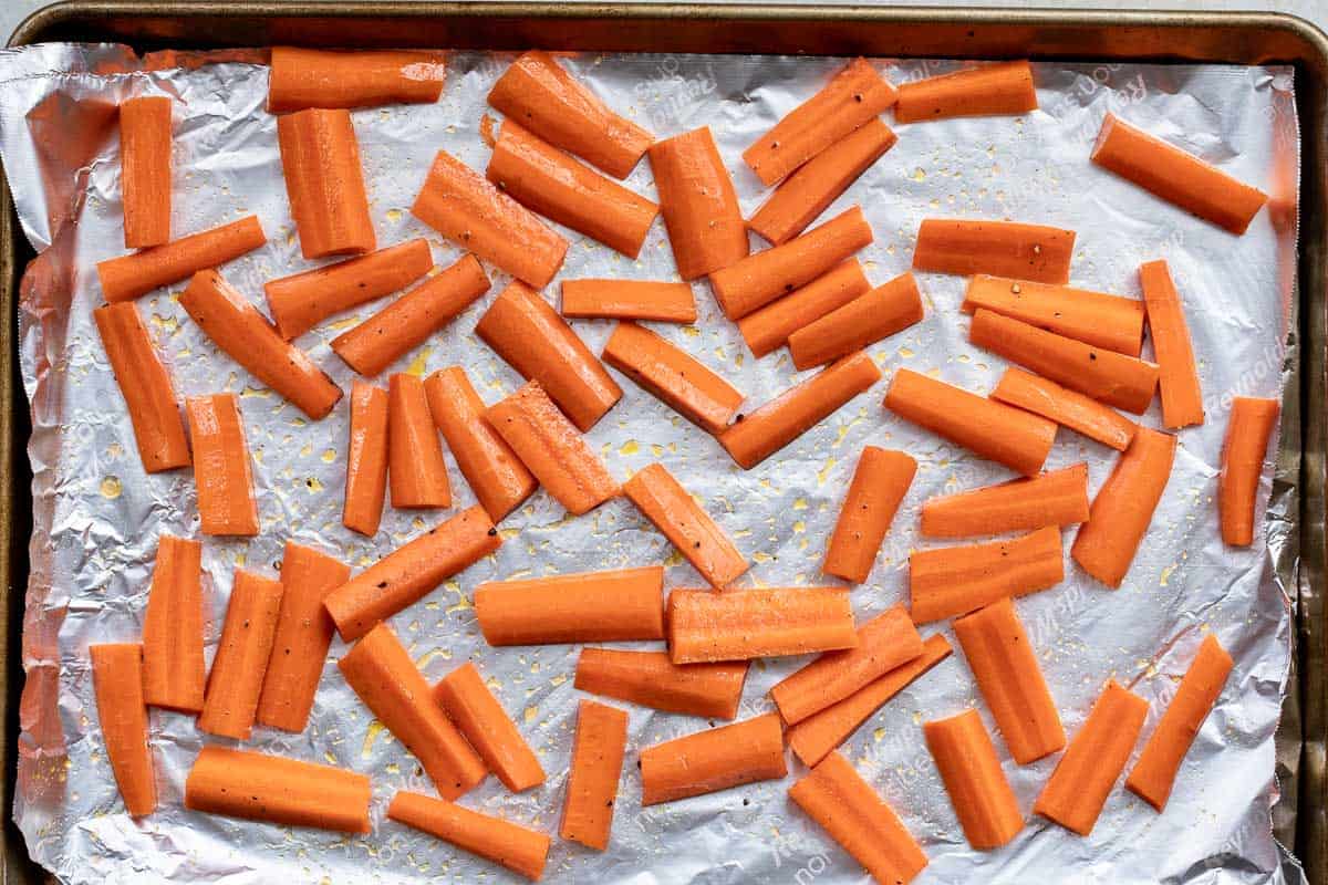 Carrot batons arranged on a foil-lined baking sheet, seasoned with salt and pepper, ready for roasting.