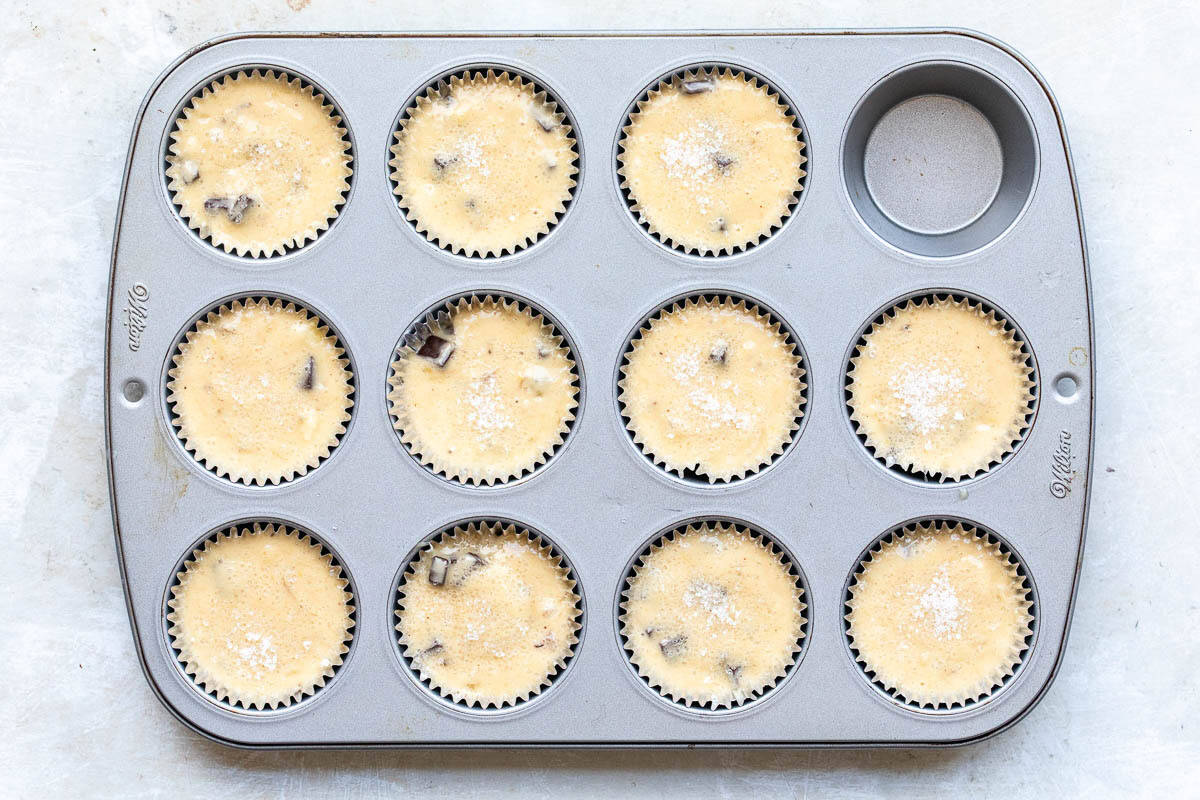 A muffin tin with eleven paper liners filled with unbaked banana muffin batter and one empty compartment, viewed from above.