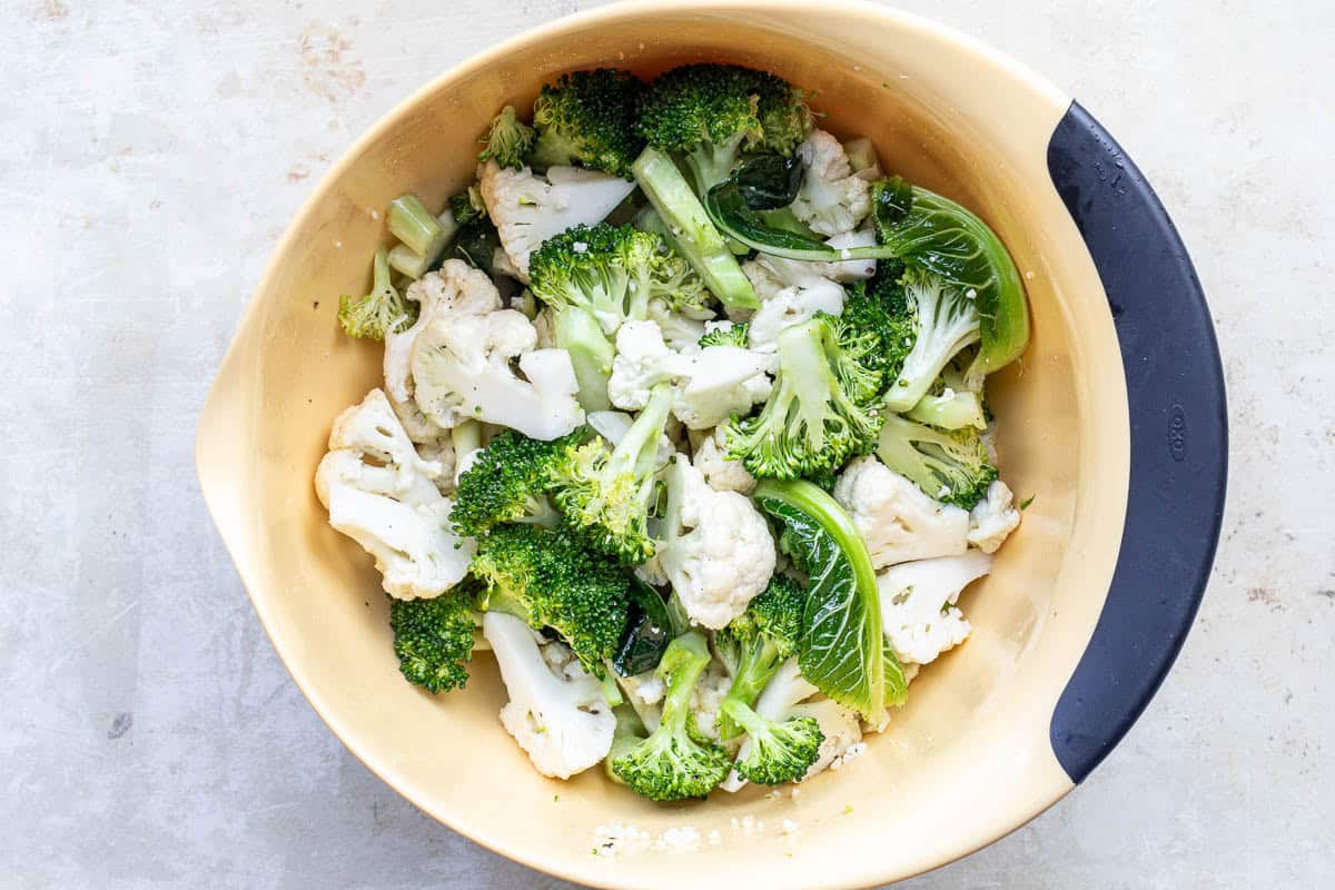 A beige mixing bowl contains raw broccoli and cauliflower florets tossed with oil, salt, and pepper.