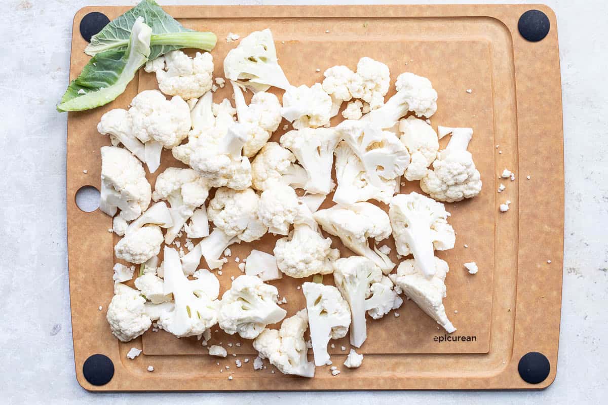 Chopped cauliflower florets and pieces are spread on a brown cutting board with a small cauliflower leaf in the corner.