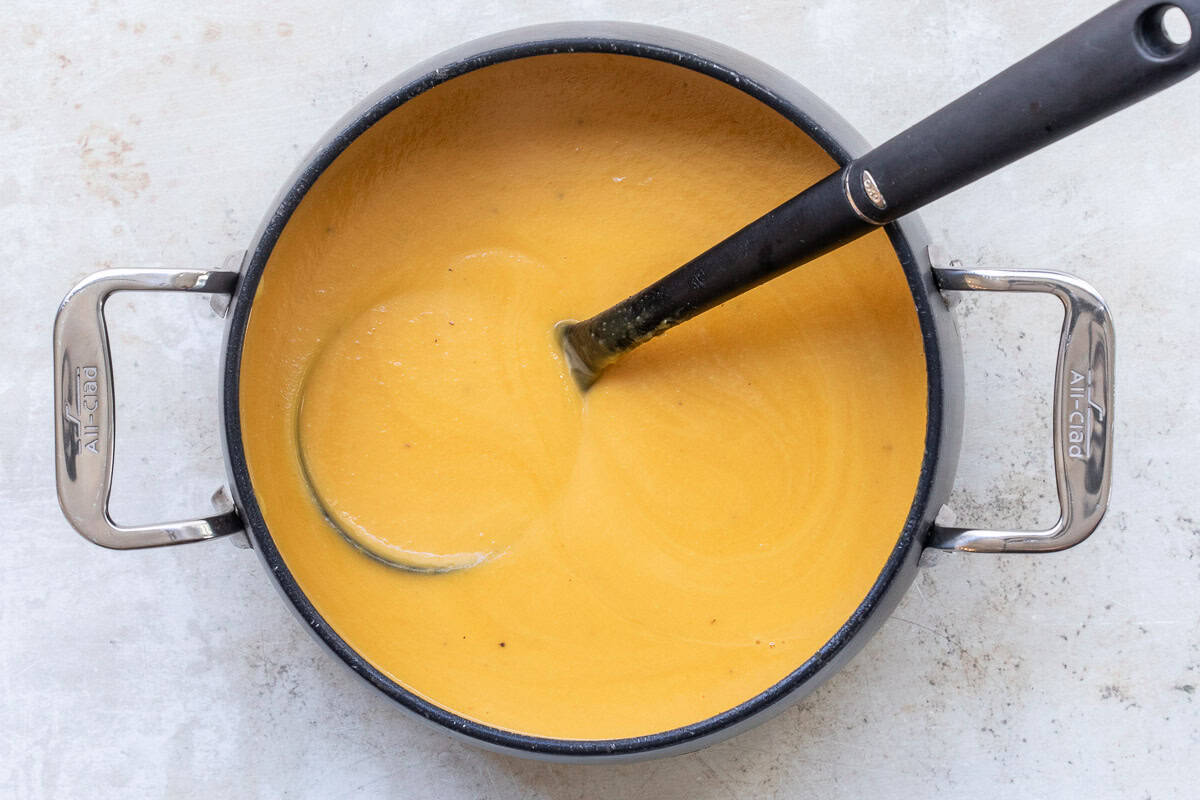A pot of creamy butternut squash soup with a black ladle resting inside, viewed from above on a light surface.
