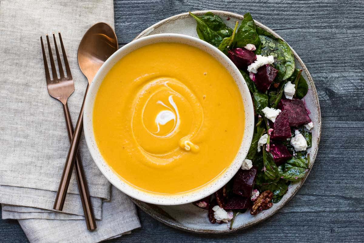 A bowl of creamy butternut squash soup with a side salad of spinach, beetroot, walnuts, and crumbled cheese, plated on a gray surface with a napkin and gold utensils.